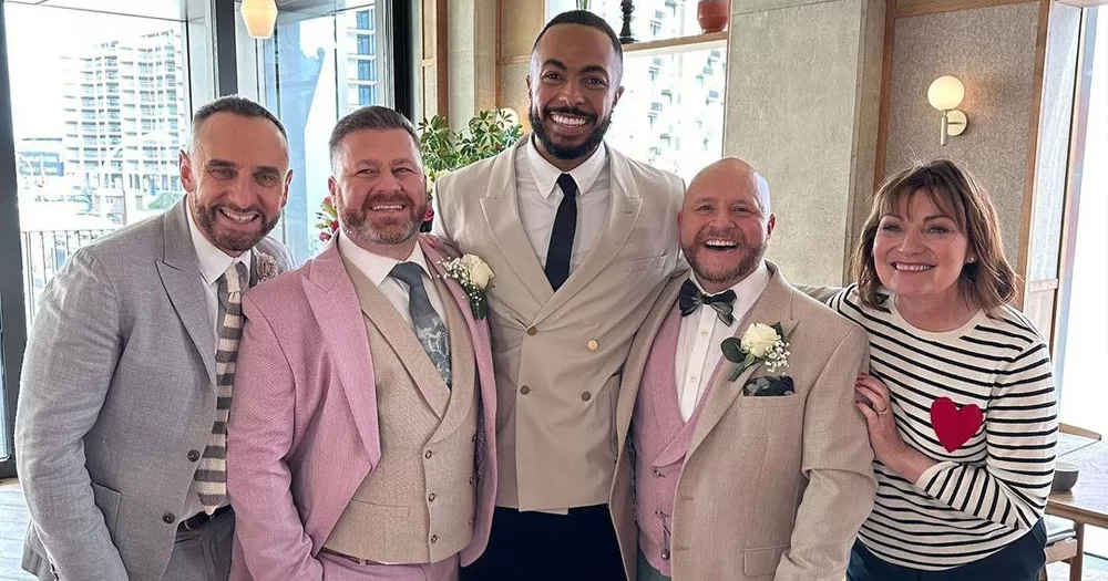 The image shows five people standing together celebrating the live wedding on the Lorraine show. In the left is a man in a grey suit, beside him is one of the grooms wearing a pink blazer and tan waistcoat. In the middle is a taller man wearing a double-breasted cream blazer with a white shirt and black tie. To his right is the second groom wearing a tan blazer and pink waistcoat and at the end is chat show host Lorraine Kelly wearing a black and white striped long-sleeved tshirt with a red heart on the right breast.