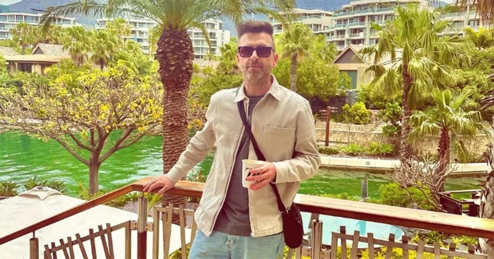 Westlife singer Mark Feehily posing for a photo in Capetown. He stands on a balcony, with palm trees visible in the background. He smiles and wears sunglasses.