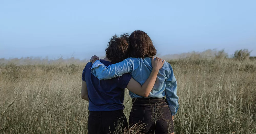 This article is about queer friendship. The image shows the backs of two people in a field, embracing as they stand side by side.