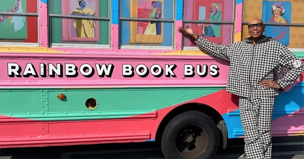 RuPaul posing with this new bookstore's Rainbow Book Bus. The bus is painted multicoloured, with RuPaul smiling to the side of it.