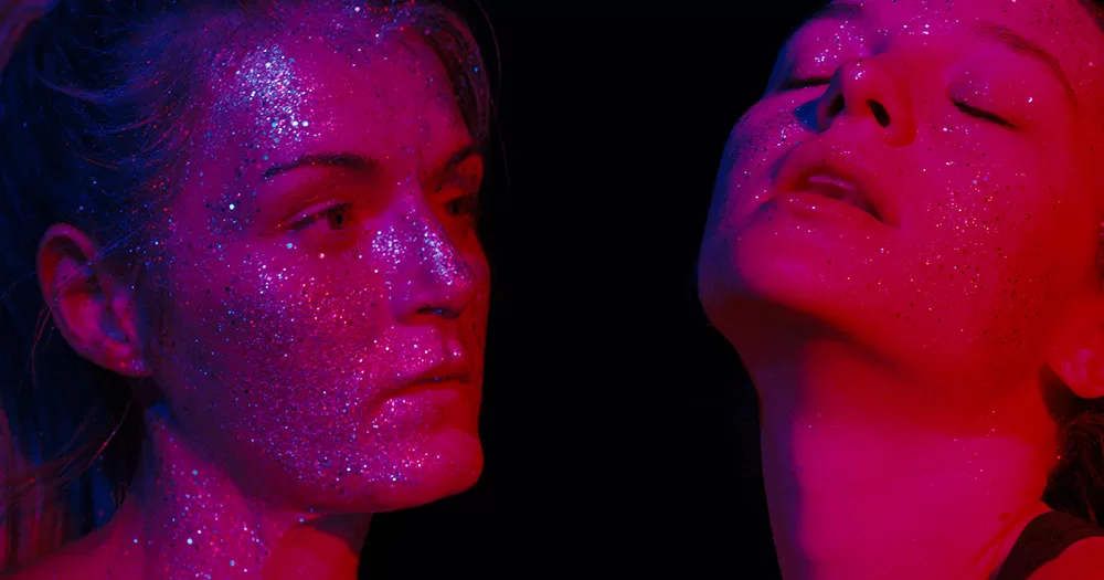 A still from Silver Haze. It shows a close up of Vicky Knight as Franky (left) with glitter all over her face in pink light, looking at the character Florence, who also has glitter on her face and closes her eyes.