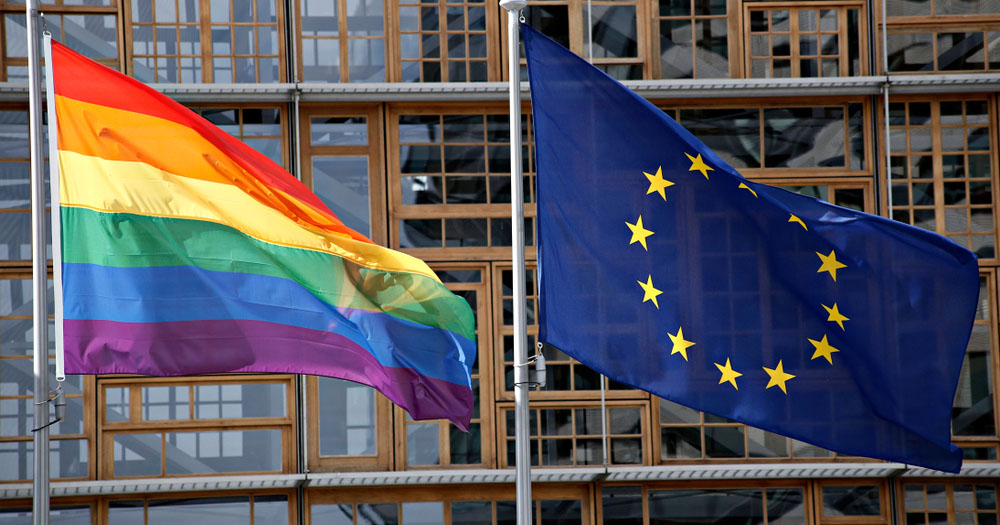 The article reports on new research into anti-gender politics in Europe. The image shows a Pride flag beside an EU flag in front of a glass and wood building.
