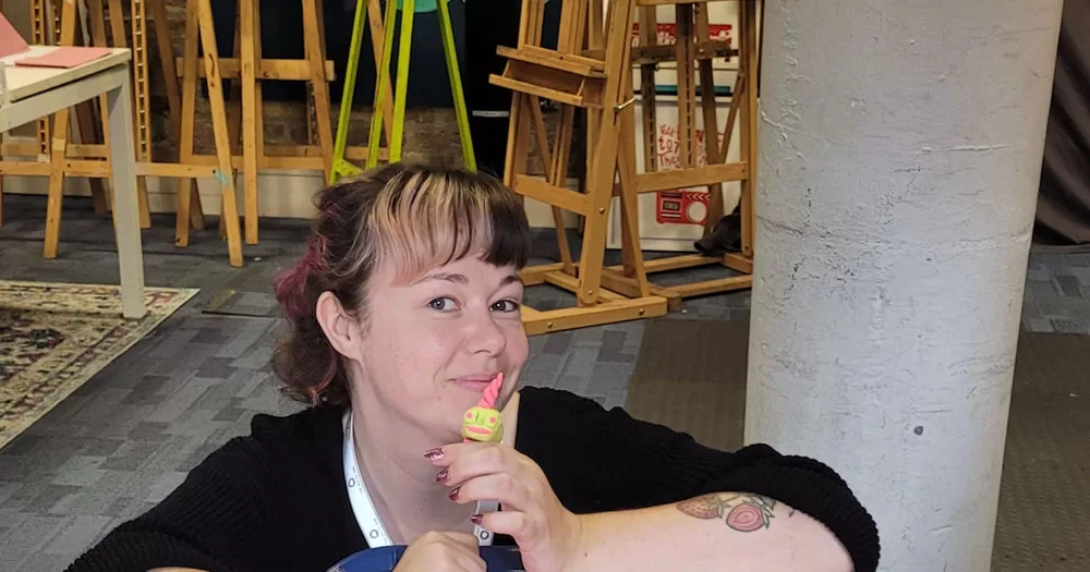 The image shows Zoe McCormack, founder of the Autistic Art Club. The photograph shows her head and shoulders leaning on a table. She is smiling and has her arms folded on the back of a chair. She is holding a yellow and pink figurine against her mouth. In the background are drawing easels.