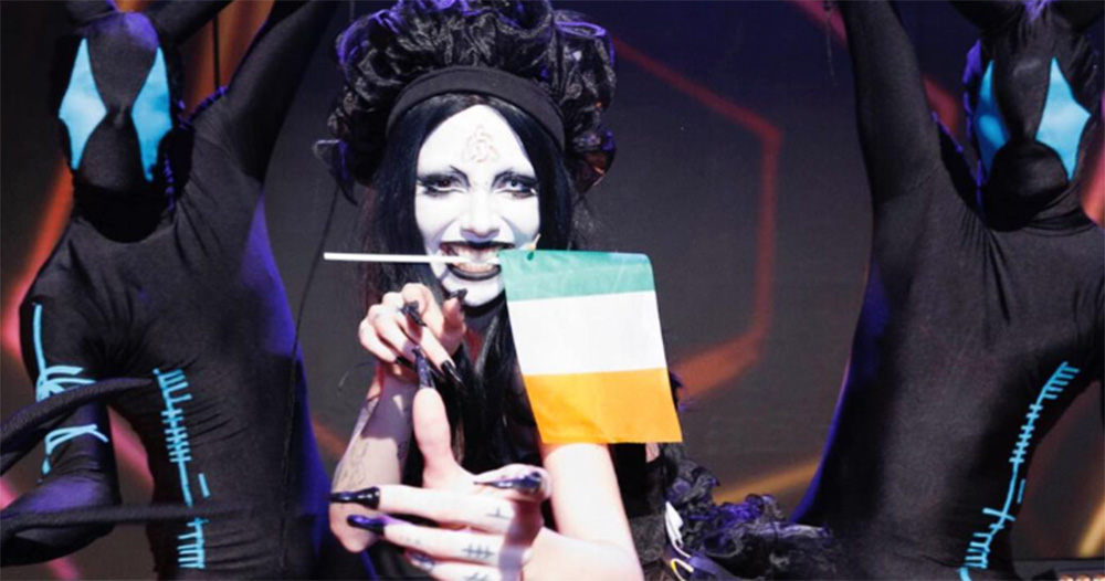 An image of Bambie Thug, who is being urged to boycott Eurovision. They pose on stage, with a small Irish flag held between their teeth as they smile. Two dancers covered in a head to two black body suit stand behind the singer on either side.