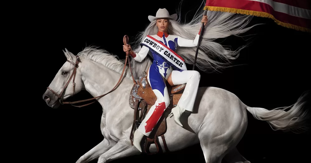 A cropped image of the cover of Beyoncé's new album Cowboy Carter. It shows Beyoncé sitting on top of a white horse, wearing a cowboy hat and rodeo outfit, and holding an American flag.
