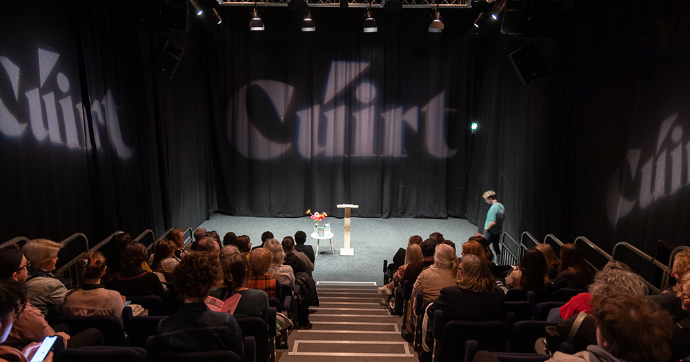 The image shows the stage at Cúirt literature festival, which features an array of queer highlights in its programme. There is an audience in the foreground looking towards the small stage, which is backdropped by black curtains with the word 'Cúirt' projected onto them.