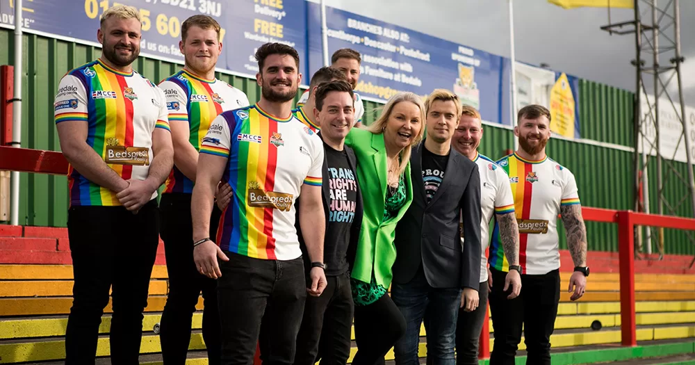 Patron India Willoughby poses with members and owners of Keighley Cougars. Players wear Pride rugby jerseys, and everyone is smiling for the camera.