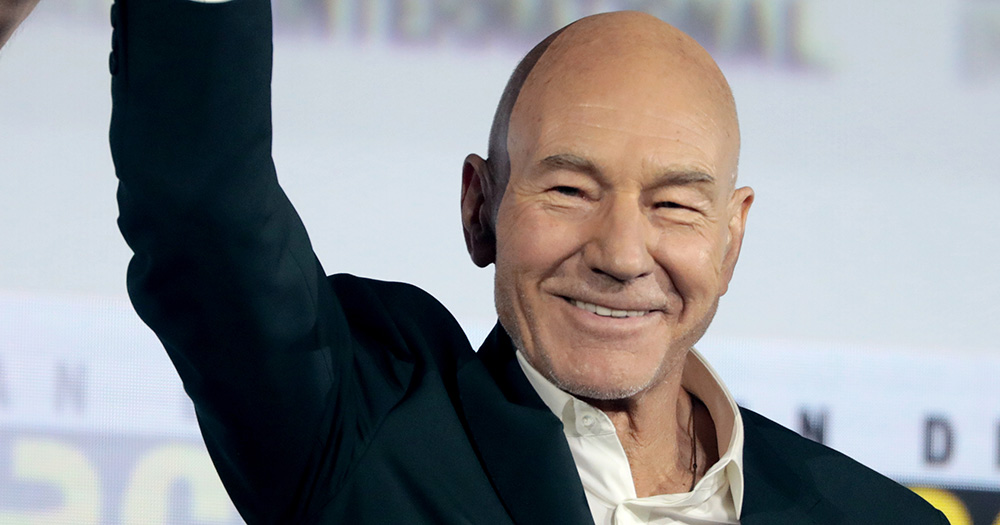A photo of gay icon Sir Patrick Stewart. He smiles, wearing a suit and waving his hand in the air.