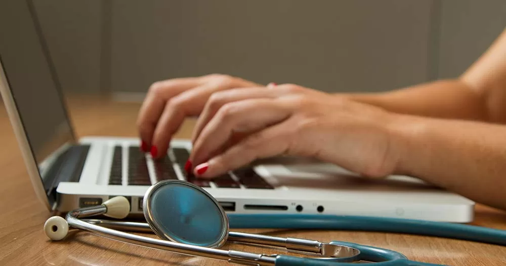 Photo of hands typing on lap top next to stethoscope representing the recent Cass Review on transgender healthcare
