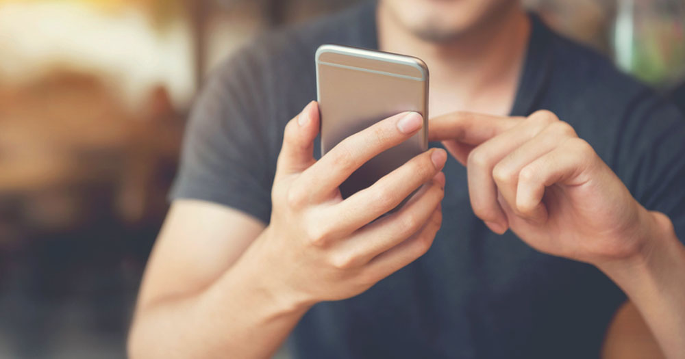 The image shows a man using a mobile phone. Male-identified users have been found to be targeted by misogynistic content on social media. In the image shows the upper body of a man. His head is cropped out. He is holding a silver phone in one hand and is using his finger to scroll with the other hand. He is wearing a navy coloured tshirt.