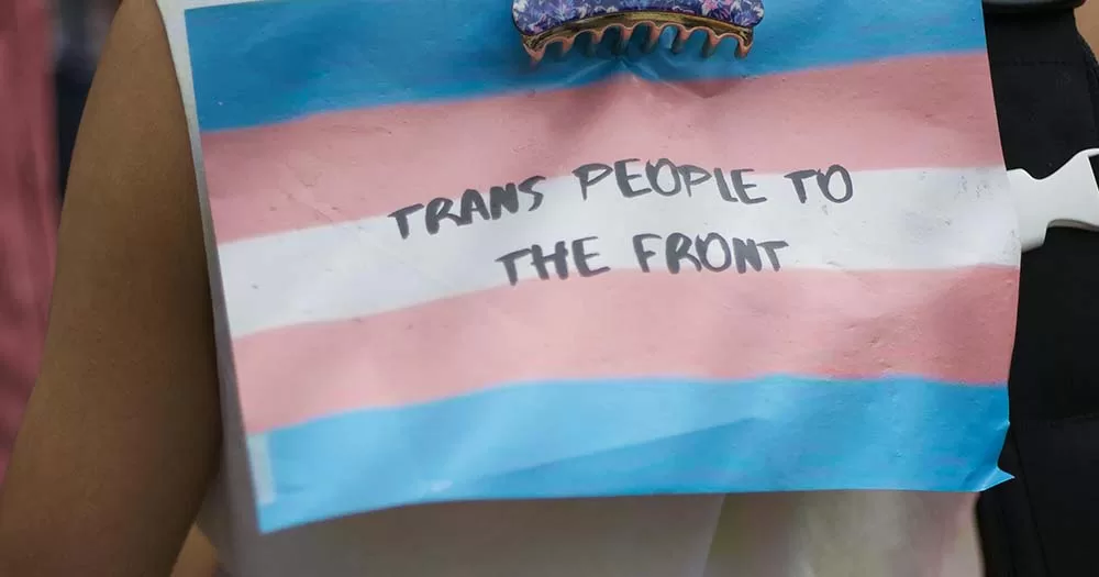 Photo of trans flag clipped to someone's shirt saying "trans people to the front", the article is about LGBTQ+ responses to the Cass review