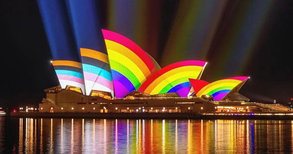 The article covers the news that new housing is to be built in Sydney for trans women. The image shows Sydney Opera House lit up in the colours of the Progressive Pride flag. The photograph is a night time shot with the sails of the Opera House reflecting rainbow colours. The two left sails show the pogressive colours of brown, black, light blue, light pink and white.
