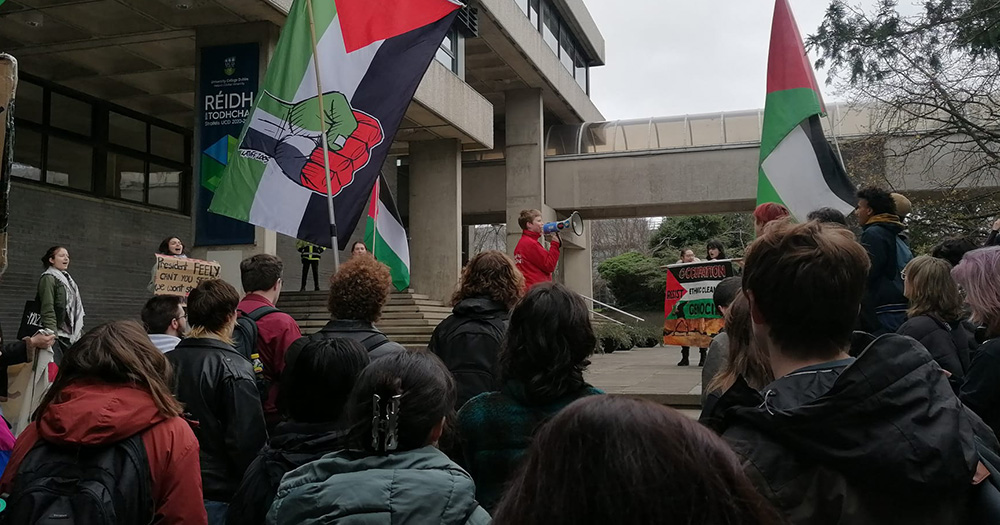 Martha Ní Riada, who was thrown out of a Nancy Pelosi event, stands in front of a crowd of people with a megaphone. Palestinian flags are being held by the crowd.