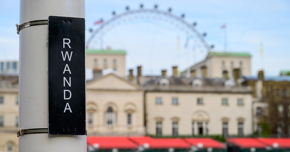 An image taken in London, UK, showing a sign post that has "RWANDA" in white text on a black background. In the background, the London Eye is visible.