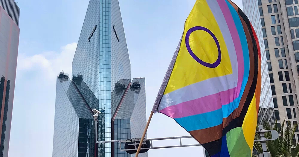 This story is about the UN adopting a resolution on intersex rights. The image shows a lard progress Pride flag flying in a stick, with a skyscraper visible in the background.
