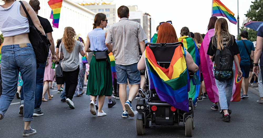 This article is about accessibility at Dublin Pride. In the photo, people marching at a Pride parade, with one of them on a wheelchair with a Pride flag on their shoulder.