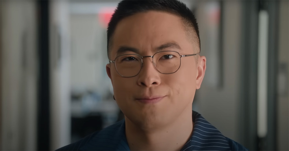 Screenshot of Bowen Yang in an SNL skit. The image is a close up of his face. He smiles slightly, has a tight haircut and is wearing glasses.