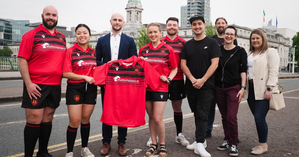 Cork Hellhounds unveiling their new rugby kit. A group of people pose outdoors for a photo, holding a red and black jersey. Some people are dressed in rugby kit, while others are dressed in regular clothes.