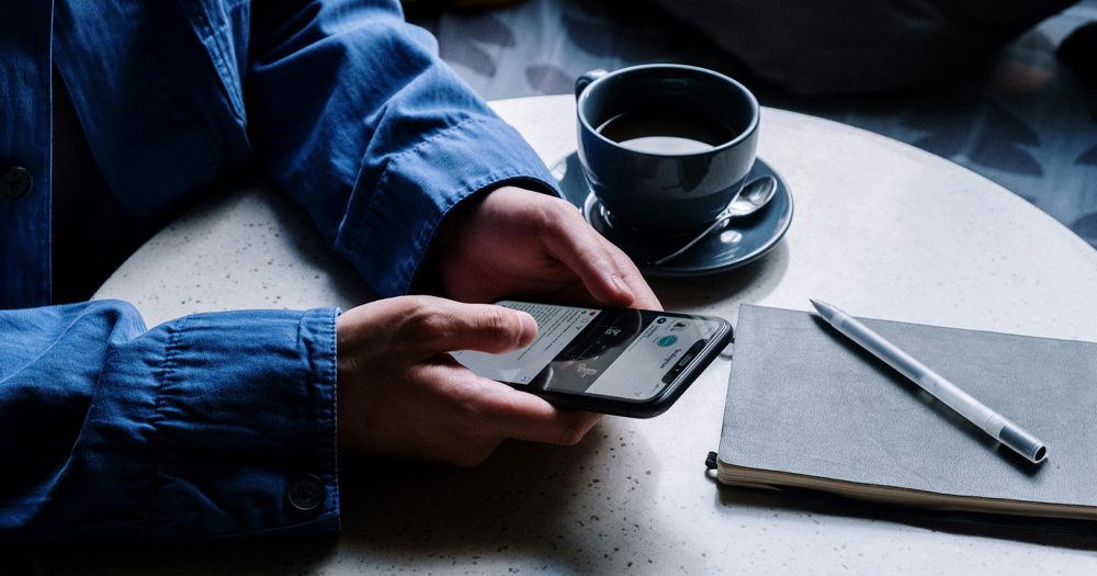 This article is about anonymous Irish accounts spreading extreme views. In the photo, the hands of a person holding a phone with a coffee and a notebook on a table.