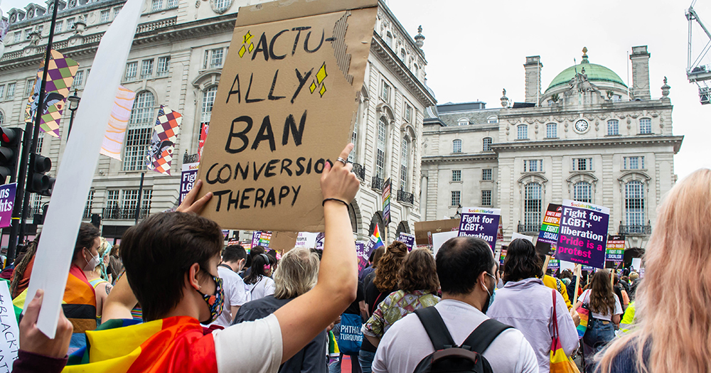 This article is about a Northern Irish study on conversion practices. In the photo, a protest with a person holding a banner that reads "Actually ban conversion therapy now"