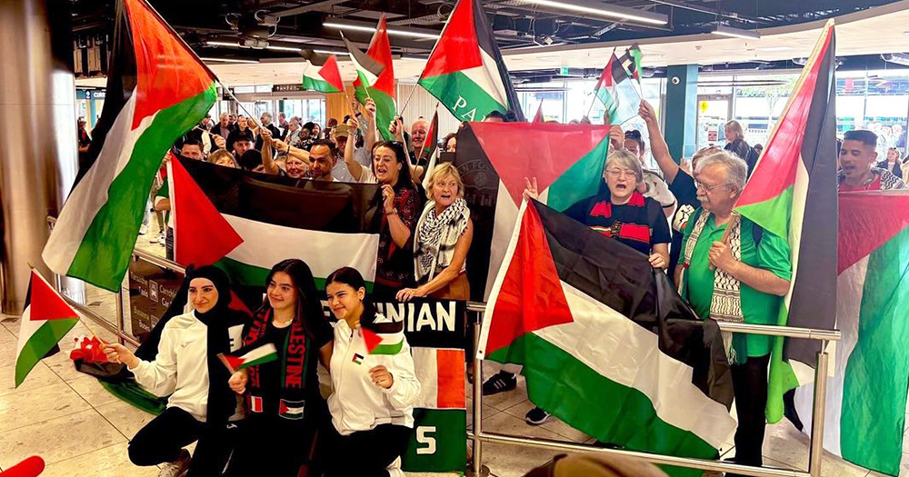 The Palestinian women's football team posing with supporters at Dublin airport, with many Palestinian flags and banners with messages of support.