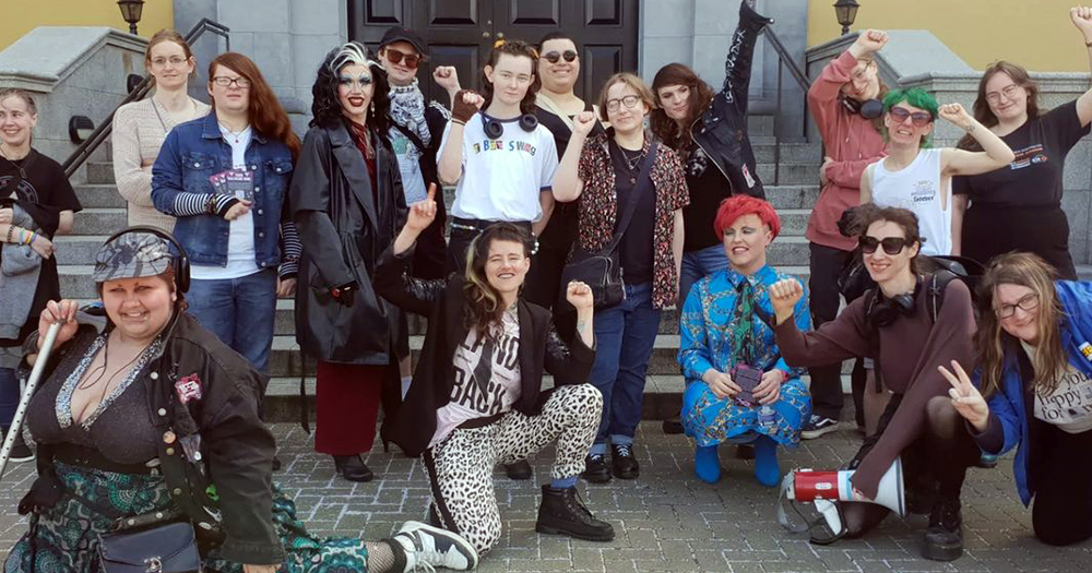 Activists staging a street theatre protest to call for better trans healthcare in Ireland. The activists are posing for a group photo, with some wearing heavy make up and colourful clothes.
