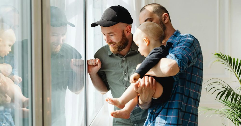 A gay couple standing in front of a window holding a baby in their arms.