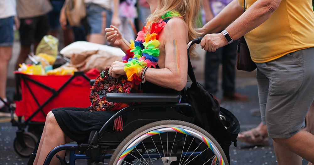 This article is about accessibility at Pride. The image shows a side view of a wheelchair user, with someone pushing them. Their wheels are decorated with rainbow designs and the person in the wheelchair wears a rainbow flower necklace and has a rainbow painted on their arm.