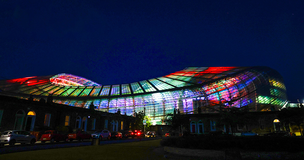 The Aviva Stadium lit up in rainbow colours for Pride. The glass stadium is showed in in a multicoloured display on a dark night.