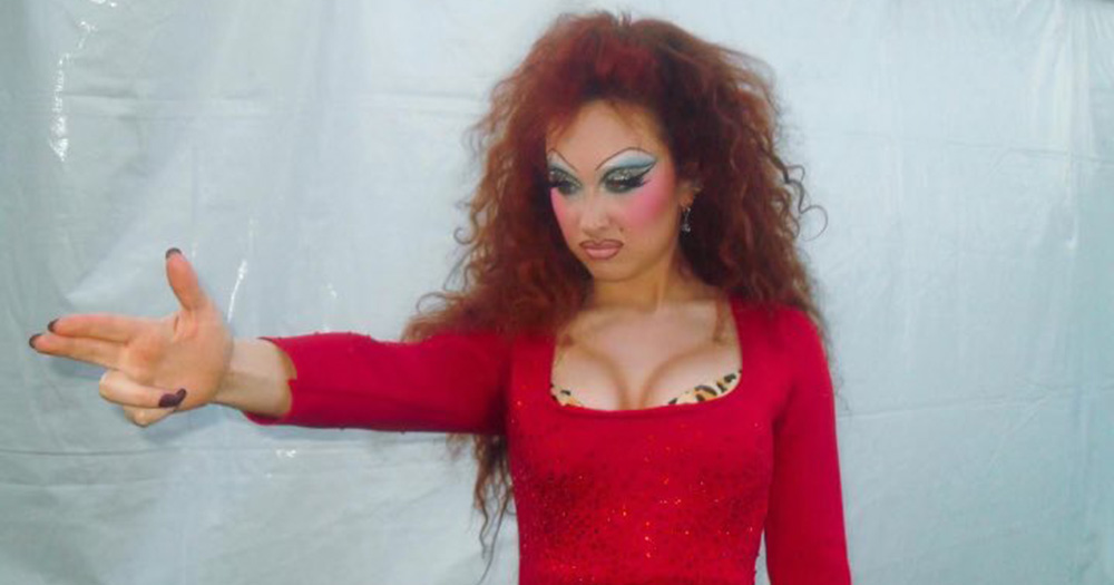 Chappell Roan dressed up as drag queen Divine. She wears a tight red dress, bold make up and messy long hair and holds her hand outstretched from her body, with finger guns.