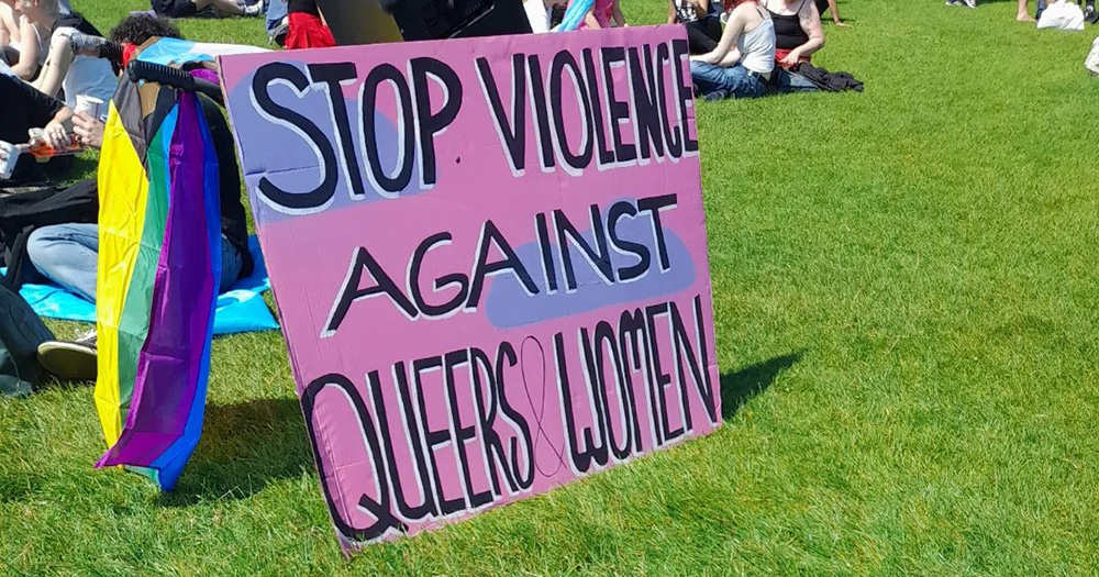 This article is about a teen who has been charged in relation to alleged incident involving gay men in Phoenix Park. The image shows a "Stop violence against queers & women" sign, perched upright on green grass.