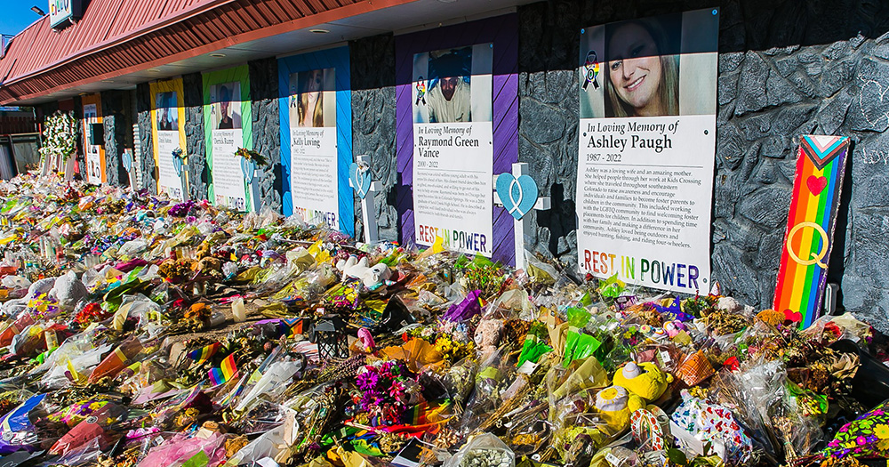 A photo of Club Q in Colorado Springs where an anti-LGBTQ+ hate crime took place in November 2022. The images shows the exterior of the venue, with the photos of victims hung up on posters on the walls. In front of the posters are piles of flowers on the ground.