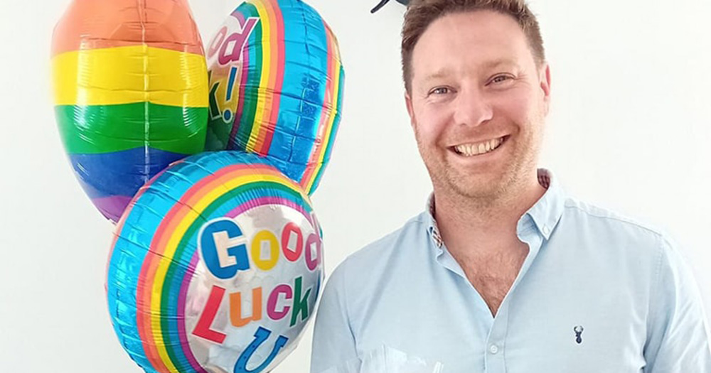 Declan Meehan photographed from the chest up, smiling and standing to the right of three balloons. There is one rainbow balloon and two colourful good luck balloons.
