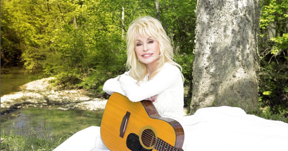 A photo of country music star Dolly Parton holding an acoustic guitar in front of a nature background.