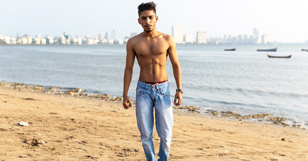 An image from Elska Mumbai. It shows a man walking shirtless on a beach wearing blue jeans.
