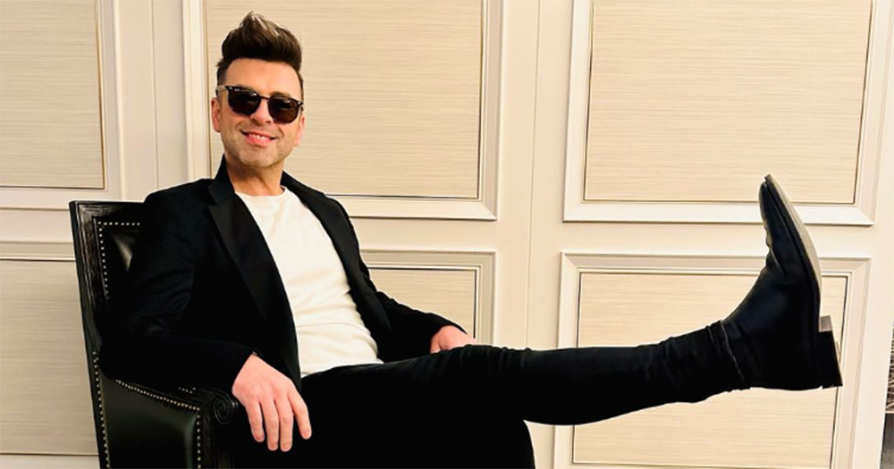 Westlife singer Mark Feehily sitting on a chair with one of his legs raise. He wears a black suit and sunglasses and smiles at the camera.