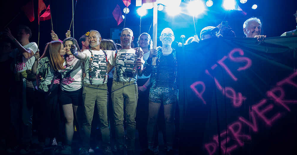 Lesbians and Gays Support the Miners stand on stage at an event, with a bright light behind them. They wear iconic group activist t-shirts and hold a banner reading "Pits & Perverts".