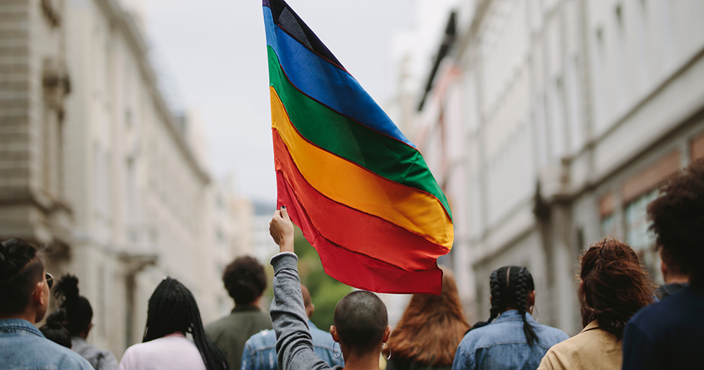 This piece about the importance of Pride in 2024. The image shows a rainbow flag being carried above a group of people walking through a city's street.