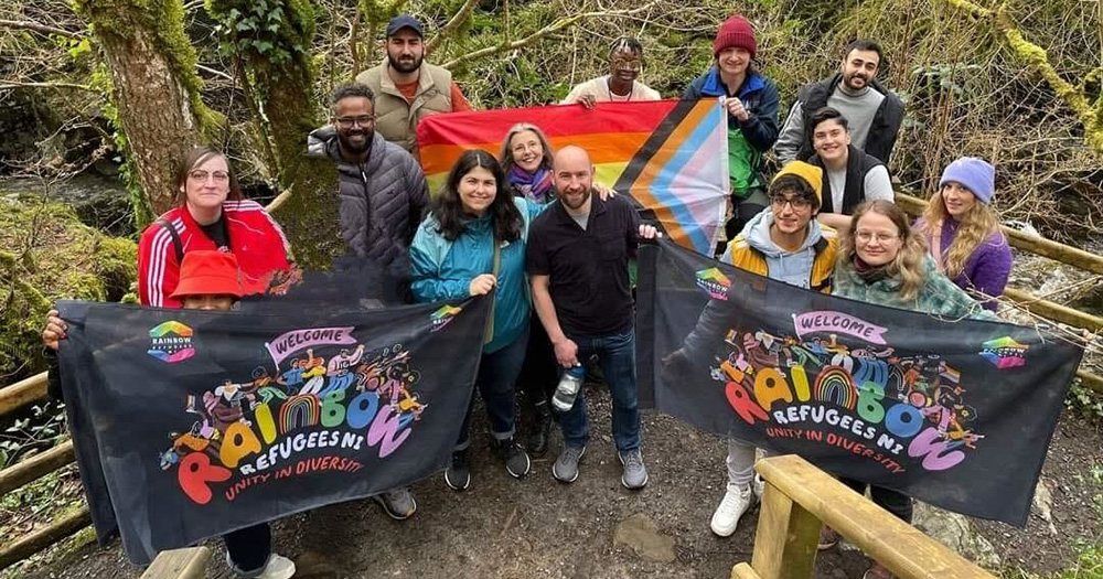 A group photo of Rainbow Refugees NI. They pose on an outdoor hike holding flags with their group logo on them.