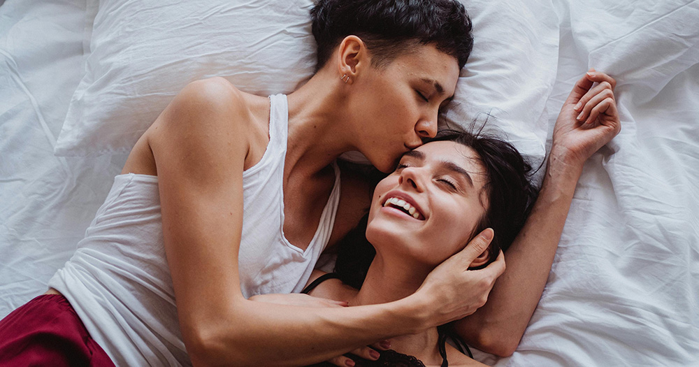 This article is about the new 'slowmance' dating trend. The image shows a queer couple in bed. The person on the left wears a white vest, has short black hair and kissed their partner on the right on the head. The person on the right has dark long hair and smiles with their eyes closed.