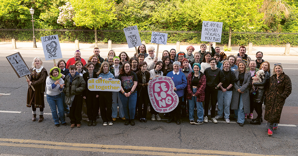 A group of young LGBTQ+ activists, standing with members of Belong To and Dublin Pride. They stand in a big group holding various signs and logos.