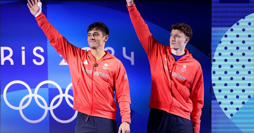 Tom Daley and diving partner Noah William after winning silver at the Paris 2024 Olympics, as they salute the crowd wearing red jumpers on a blue background.