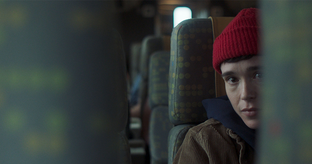 A scene in film 'Close To You,' Actor Elliot Page sitting inside a large bus wearing a red beanie, blue hoodie and brown jacket; the image looks ahead to him from in-between two bus seats.