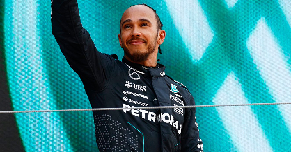 Image of Lewis Hamilton, who praised Ralf Schumacher for coming out. Lewis is photographed from the stomach up in his racing uniform, and raises his right arm in the air. He smiles and looks into the distance.
