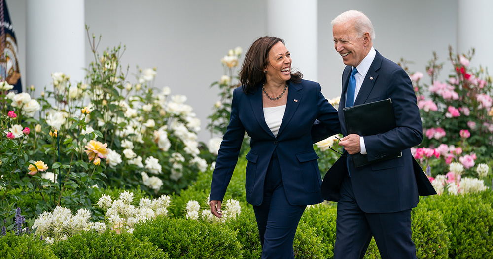 An image of Kamala Harris and Joe Biden walking and laughing side by side outdoors. They both wear navy suits, and flowers and bushes can be seen in the background.