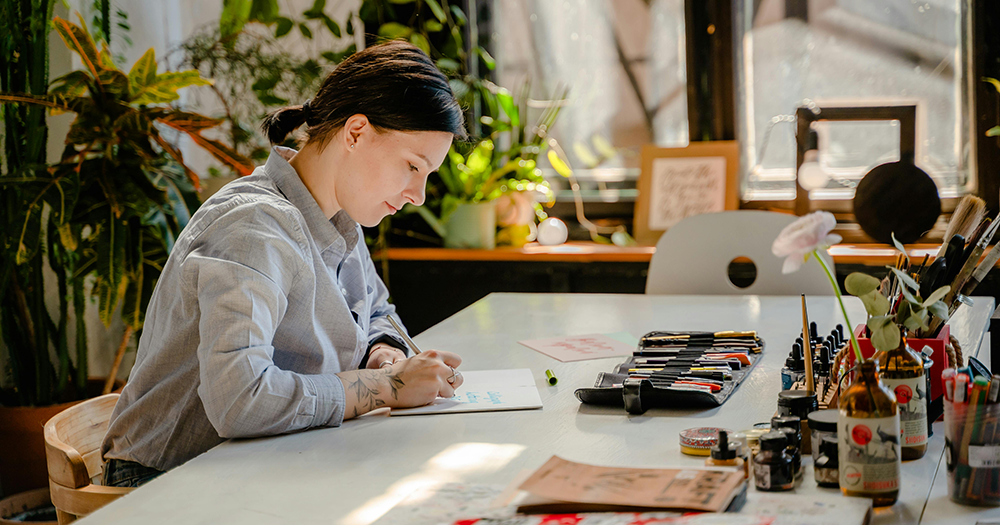 This article is about LGBTQ+ artists and arts workers facing workplace discrimination. The image shows a woman sitting at a desk with pens, pencils and markers in front of her. She draws on a piece of paper, and lots of plants can be seen in the background.