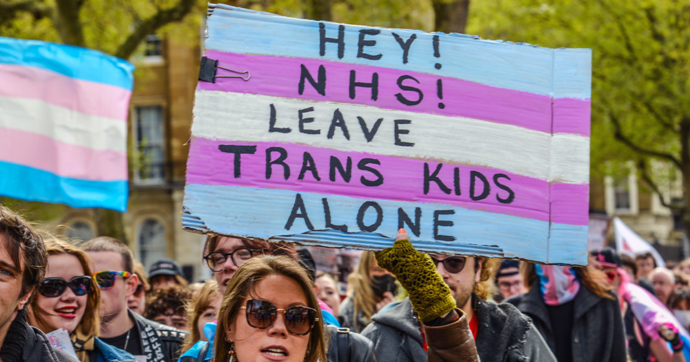This article is about a crowdfunder launched by Mermaids. In the photo, some protesters holding trans flags and a sign that reads "Hey NHS, leave trasn kids alone".