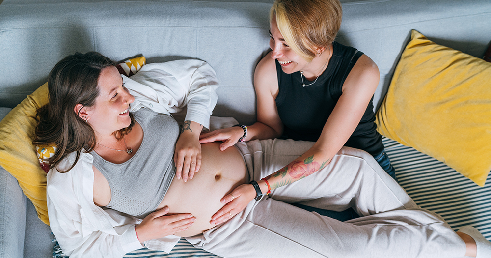 This article is about a webinar hosted by Thérapie Fertility. In the photo, two women laying on a grey sofa and smiling at each other. One of the women is pregnant.