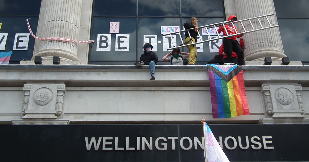 Protesters gather on the ledge of the NHS building with signs of support for trans youth.