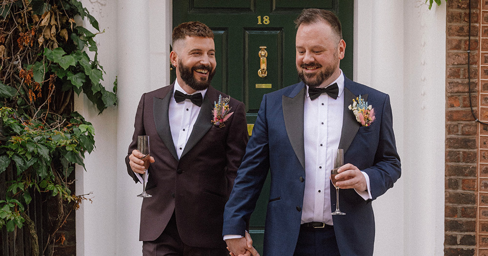 Stefano Pappalardo and husband Aaron Healy in Best Menswear Made To Measure suits. The pair hold hands and smile holding champagne glasses with a green door in the background.